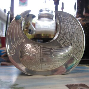 May include: A silver-toned metal bird-shaped sculpture with intricate etched details. The bird has a turquoise-colored stone eye and a detailed wing design. The sculpture is likely a decorative item or a piece of art.