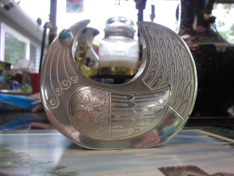 May include: A silver-toned metal bird-shaped sculpture with intricate etched details. The bird has a turquoise-colored stone eye and a detailed wing design. The sculpture is likely a decorative item or a piece of art.
