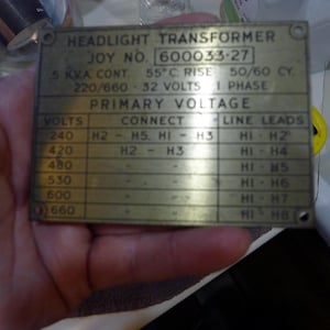 May include: A rectangular metal plate with the text "HEADLIGHT TRANSFORMER" and "JOY NO. 600033-27". The plate includes technical specifications such as voltage, phase, and connection details, with a grid of data. The plate is held by a hand.