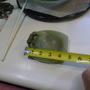 May include: Green ashtray with a bronze-colored dog figurine on the side. The ashtray is approximately 4 inches long. The ashtray is made of a smooth, polished material. The ashtray is a vintage item.