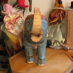 Puede incluir: Una figurita de elefante de madera pintada de azul y dorado con la trompa levantada y detalles decorativos. El elefante est&aacute; de pie sobre una superficie de madera.