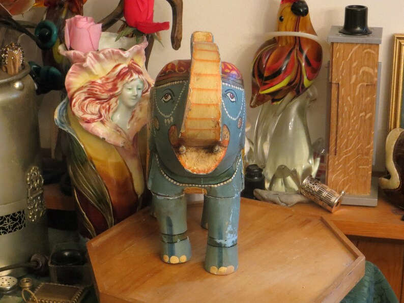 Puede incluir: Una figurita de elefante de madera pintada de azul y dorado con la trompa levantada y detalles decorativos. El elefante est&aacute; de pie sobre una superficie de madera.