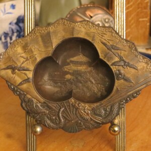 May include: A small, ornate, gold-coloured metal trinket dish with a three-leaf design. The dish features a raised, detailed image of a Japanese pagoda and surrounding landscape.