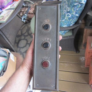 May include: A rectangular, antique elevator control panel with the words "UP", "DOWN", and "BELL" above three circular buttons. The buttons are black and red, set against a weathered, metallic background.