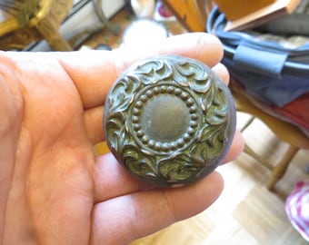 May include: A round, ornate, brass door knob with a detailed floral design. The knob is a dark green color with a circular center.