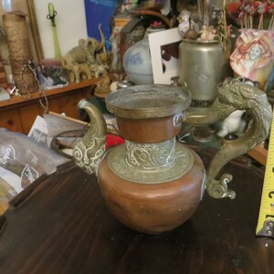May include: A copper teapot with a dragon-shaped handle and intricate brass detailing. The teapot has a round body and a wide spout. The handle is made of brass and is shaped like a dragon's head. The teapot is decorated with brass accents and has a brass lid.