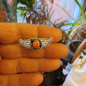 May include: A white plastic pin with blue wings and a red and yellow McDonald's logo. The pin says "McDonald's Racing Team".