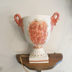 May include: A white ceramic lamp with a pink cherub design and gold accents. The lamp has two handles and a square base.