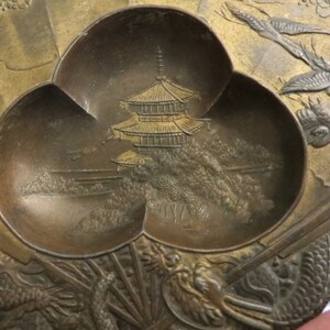 May include: A small, three-sectioned, brass tray with a raised, embossed design. The centre section features a pagoda and trees, while the other sections have stylised birds in flight.