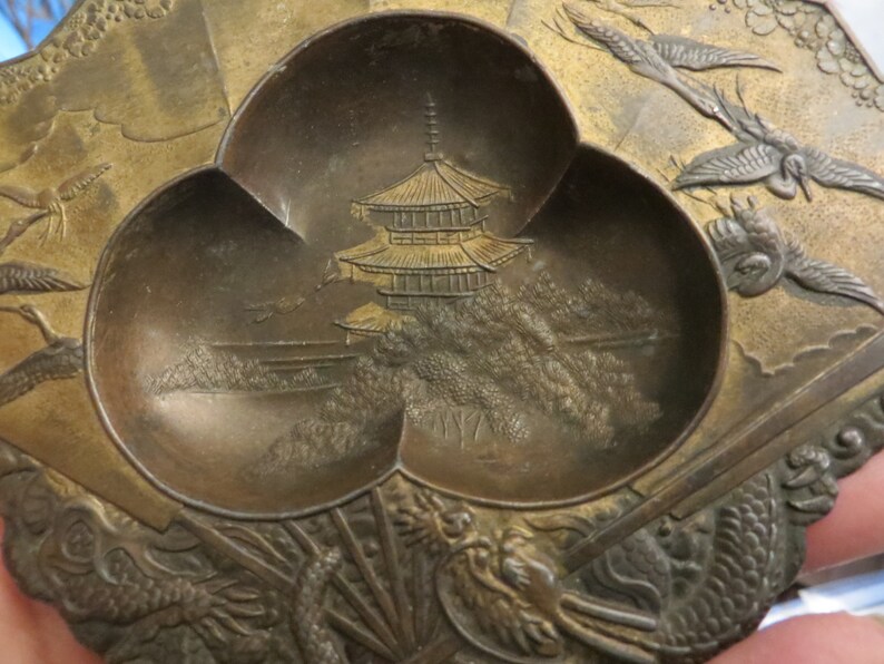 May include: A small, three-sectioned, brass tray with a raised, embossed design. The centre section features a pagoda and trees, while the other sections have stylised birds in flight.