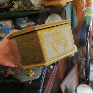 May include: A wooden box with a gold and white painted finish. The box has a decorative design on the front and three gold feet.