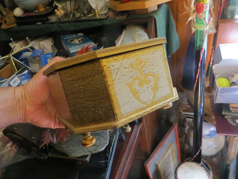 May include: A wooden box with a gold and white painted finish. The box has a decorative design on the front and three gold feet.