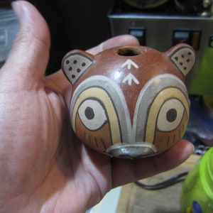 May include: A hand-painted ceramic animal figurine in the shape of a raccoon. The figurine is brown with white and black accents. The raccoon has a small hole on the top of its head.