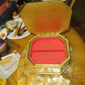 May include: A gold and white octagonal jewellery box with a red velvet interior. The box is open and the lid is propped up. The box has a decorative design on the outside.