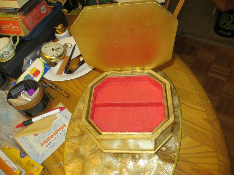 May include: A gold and white octagonal jewellery box with a red velvet interior. The box is open and the lid is propped up. The box has a decorative design on the outside.