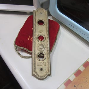 May include: A gold-colored metal elevator button plate with the words "UP" and "DOWN" and a decorative design.