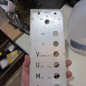 May include: A weathered metal elevator control panel with the name "DIMAK" at the top. It features buttons labeled "OFF," "ON," and "STOP," along with floor indicators like "VERANDAH DECK" and "MAIN DECK."