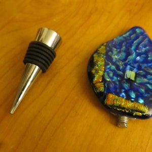 May include: A silver bottle stopper with a black rubber ring and a blue and gold glass bottle stopper with a metallic finish.