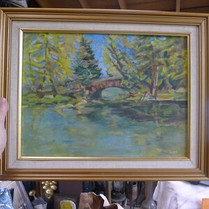 May include: Oil painting of a park scene with a stone bridge over a lake. The artwork features a gold-colored frame and depicts trees with green and yellow foliage, a blue sky, and water reflections. The painting is signed in the lower right corner.