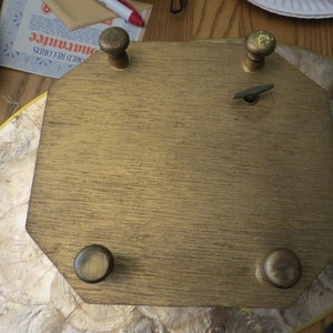 May include: A gold-coloured wooden music box with a winding key and four decorative knobs. The music box is on a light brown surface.