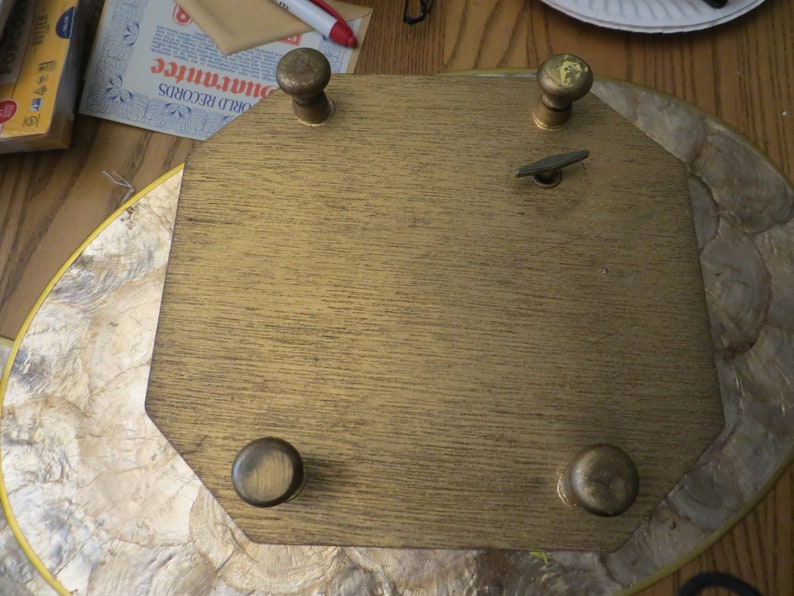 May include: A gold-coloured wooden music box with a winding key and four decorative knobs. The music box is on a light brown surface.