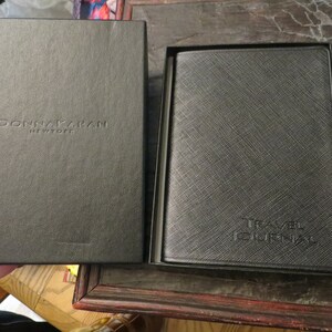 May include: Black travel journal in a matching black box. The journal cover has a textured pattern and the words "TRAVEL JOURNAL" embossed on the front. The box is embossed with the words "DONNA KARAN NEW YORK".