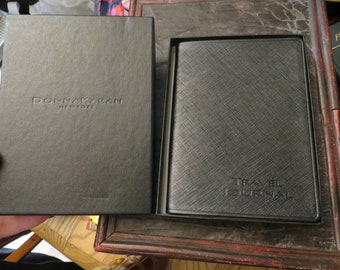 May include: Black travel journal in a matching black box. The journal cover has a textured pattern and the words "TRAVEL JOURNAL" embossed on the front. The box is embossed with the words "DONNA KARAN NEW YORK".