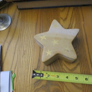 May include: A beige star-shaped trinket box with gold stars painted on it. The box is sitting on a wooden surface.