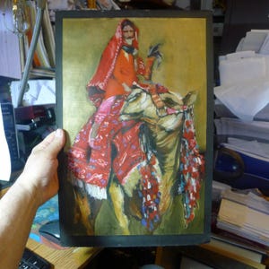 May include: An original painting depicting a person riding a camel. The person is wearing a red head covering and orange clothing. The camel is adorned with colorful decorations. The background is gold.