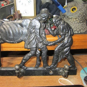 May include: A black metal wall hook featuring a horse and two figures in period clothing. The figures appear to be interacting with the horse. The hook has three decorative knobs for hanging items. The overall theme is vintage decor.