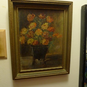 May include: Oil painting of a floral arrangement in a dark vase, set against a muted background. The flowers are a mix of orange and yellow hues, with visible brushstrokes. The painting is framed in a gold-toned, ornate frame.
