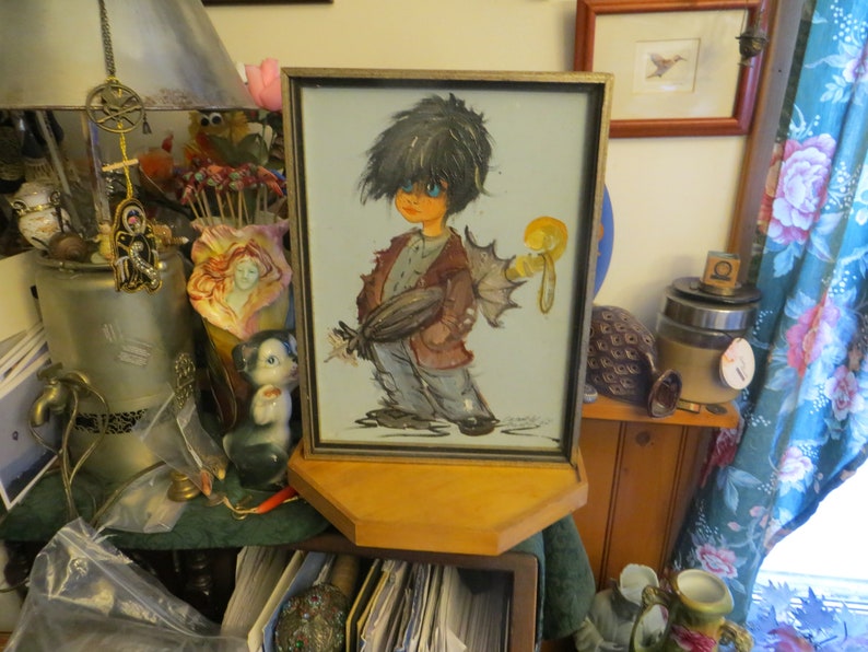 May include: A framed painting of a cartoon character with black hair, blue eyes, and a brown jacket. The character is holding a yellow object in their right hand. The painting is set on a wooden shelf.