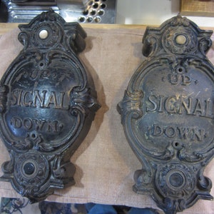May include: Two antique black metal elevator signal plates with ornate detailing. Each plate has a round button and the text "UP SIGNAL DOWN".