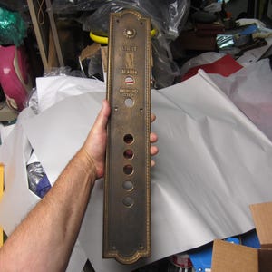 May include: A vintage elevator control panel with a bronze finish. The panel has the words "LIGHT", "ALARM", and "EMERGENCY STOP". It features several circular buttons and a decorative border. The panel is held by a person.