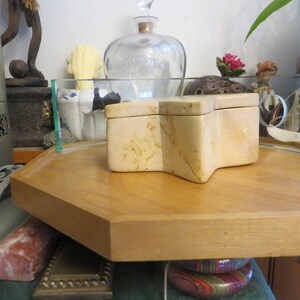 May include: A star-shaped, beige stone box with a lid, sitting on a wooden tray. The box is made of natural stone with a marbled pattern.