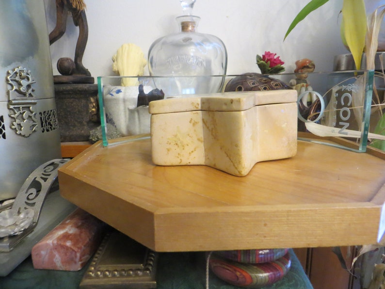 May include: A star-shaped, beige stone box with a lid, sitting on a wooden tray. The box is made of natural stone with a marbled pattern.