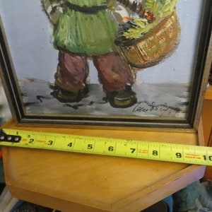 May include: A framed painting of a person carrying a basket of flowers. The painting is signed "Lucy Dolly" in the bottom right corner. The frame is black with a gold trim.