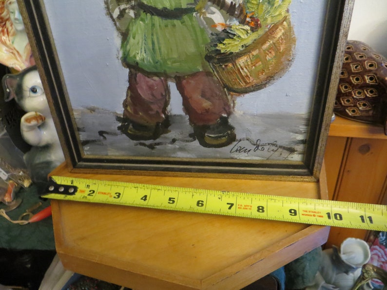 May include: A framed painting of a person carrying a basket of flowers. The painting is signed "Lucy Dolly" in the bottom right corner. The frame is black with a gold trim.