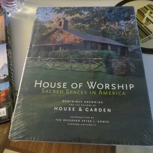 May include: A green hardcover book titled "House of Worship: Sacred Spaces in America" by Dominique Browning and the editors of House & Garden. The book features a photograph of a wooden building with a fence and a stone wall.