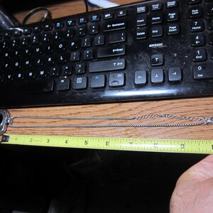 Pu&ograve; includere: Una collana color argento con un medaglione decorativo, lunga circa 25 cm, &egrave; mostrata su una superficie di legno. Una tastiera nera &egrave; sullo sfondo. La collana viene misurata con un metro a nastro giallo.