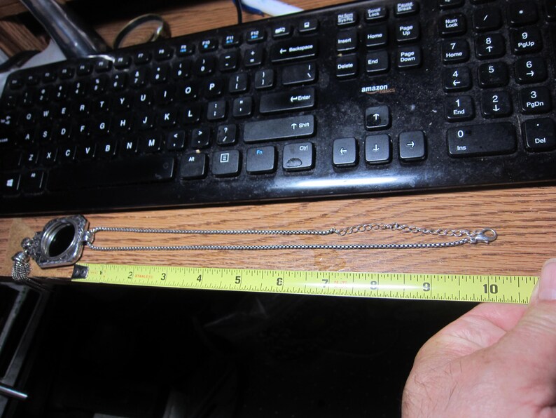 Pu&ograve; includere: Una collana color argento con un medaglione decorativo, lunga circa 25 cm, &egrave; mostrata su una superficie di legno. Una tastiera nera &egrave; sullo sfondo. La collana viene misurata con un metro a nastro giallo.