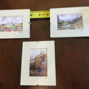 May include: Three framed watercolor paintings, each with a different scene. The first painting shows a beach scene with people walking on the sand. The second painting shows a rural scene with a farmhouse and barns. The third painting shows a moose in a forest setting.