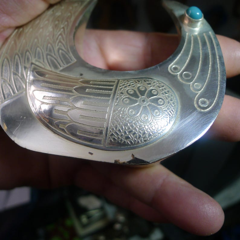 May include: A silver-toned pendant in a crescent shape, featuring engraved bird-like details and a turquoise stone accent. The pendant has intricate patterns and a polished finish, showcasing a vintage or handcrafted aesthetic.