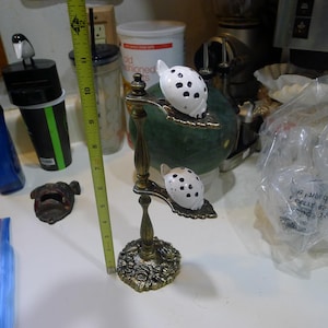 Op de afbeelding: Een messingkleurige metalen standaard met een bloemvormige voet en twee bladvormige plateaus, elk met een wit keramieken lieveheersbeestje met zwarte stippen. Een geel meetlint ernaast toont een totale hoogte van ongeveer 28 cm.