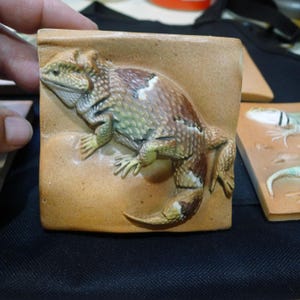 May include: A square ceramic tile featuring a three-dimensional relief of a horned lizard. The lizard is painted in shades of green, brown, and white, with textured scales and a prominent tail, set against a smooth, tan-colored background surface.