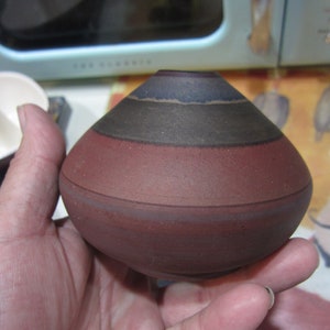 May include: A small, round, brown ceramic vase with three bands of dark brown, black, and light brown glaze.