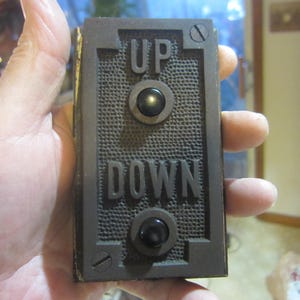 May include: A rectangular, dark brown elevator call button with the words "UP" and "DOWN" in raised letters. It has two black, round buttons and a textured background. The button is held in a hand.