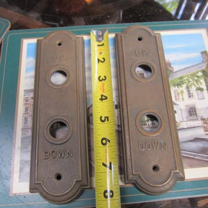 May include: Two vintage bronze elevator call plates. One plate reads "UP" and the other reads "DOWN". Each plate has a circular button and is approximately 6 inches long. A yellow measuring tape is placed between the plates.
