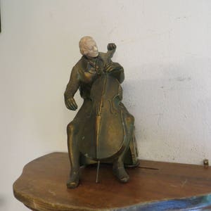 May include: A bronze sculpture of a person playing a cello. The sculpture is on a wooden shelf.