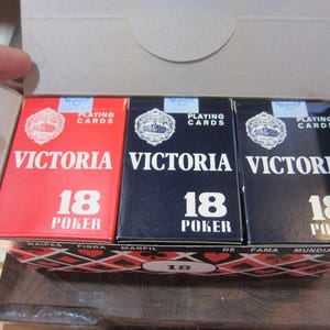May include: A box of Victoria playing cards, with three decks visible. The decks are red and navy blue, with the word "VICTORIA" and "18 POKER" printed on the front. The box is open, revealing the cards inside.